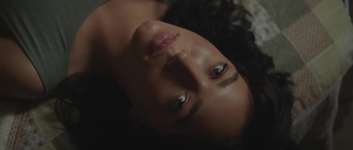 A young woman resting on a bed raises her eyes to look into the camera