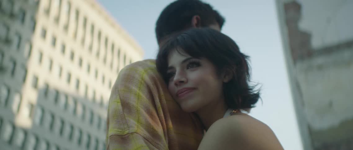 Woman wearing halter top hugs man with a smile surrounded by apartment buildings under a clear blue sky