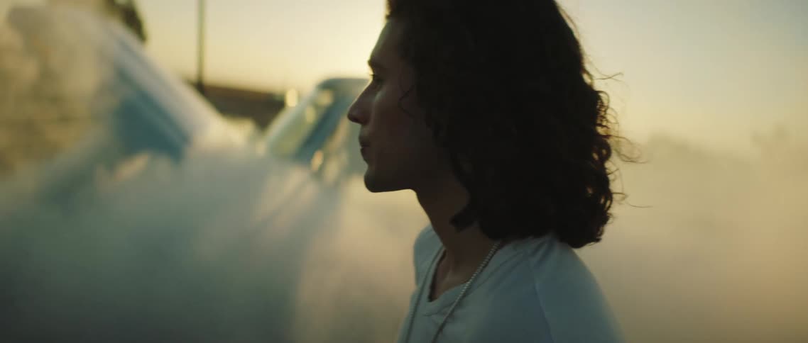 Young long haired man standing on a rural road next to a broke smoking car at sunset