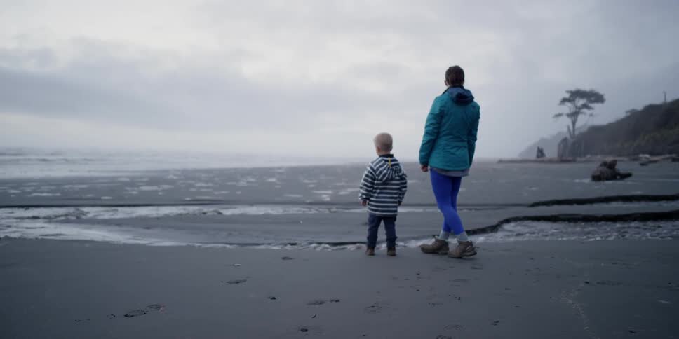 A woman and toddler boy stand on a beach watching the water