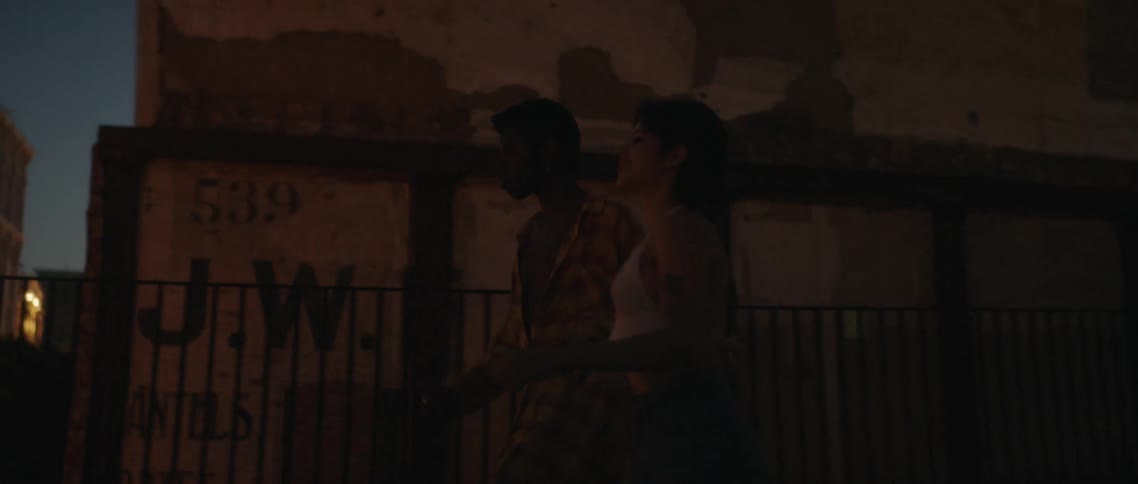 A young man and woman holding hands run to the edge of a rooftop and look down over the edge in a metropolis at night
