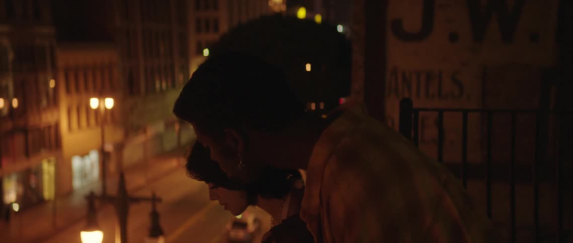 A man and woman hold hands while running to the edge of a rooftop to look at the street below at night