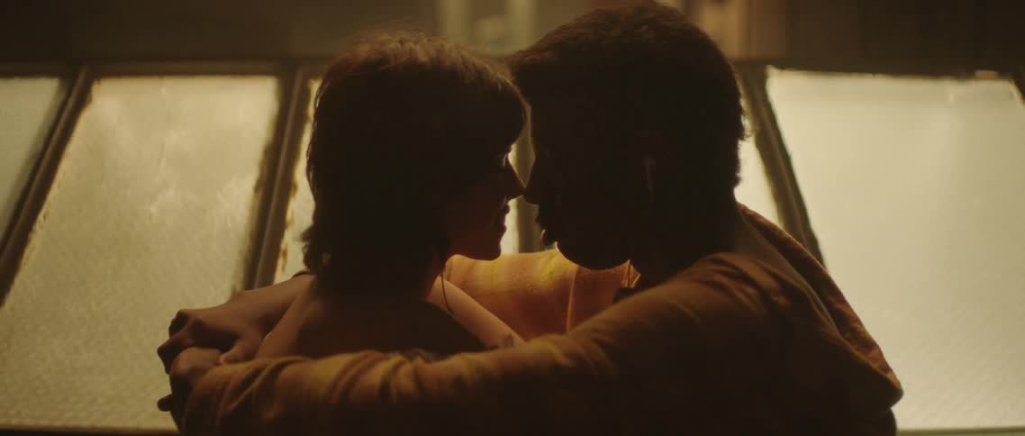 A man and woman sit on a rooftop near a skylight completely wrapped around each other face to face