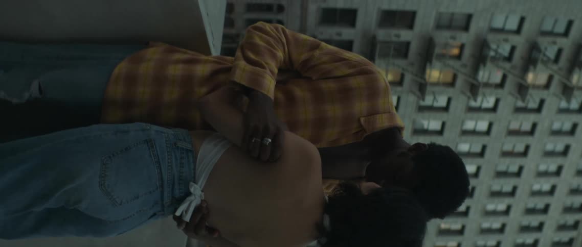 A teenage boy and girl stand on a roof top in a city and kiss