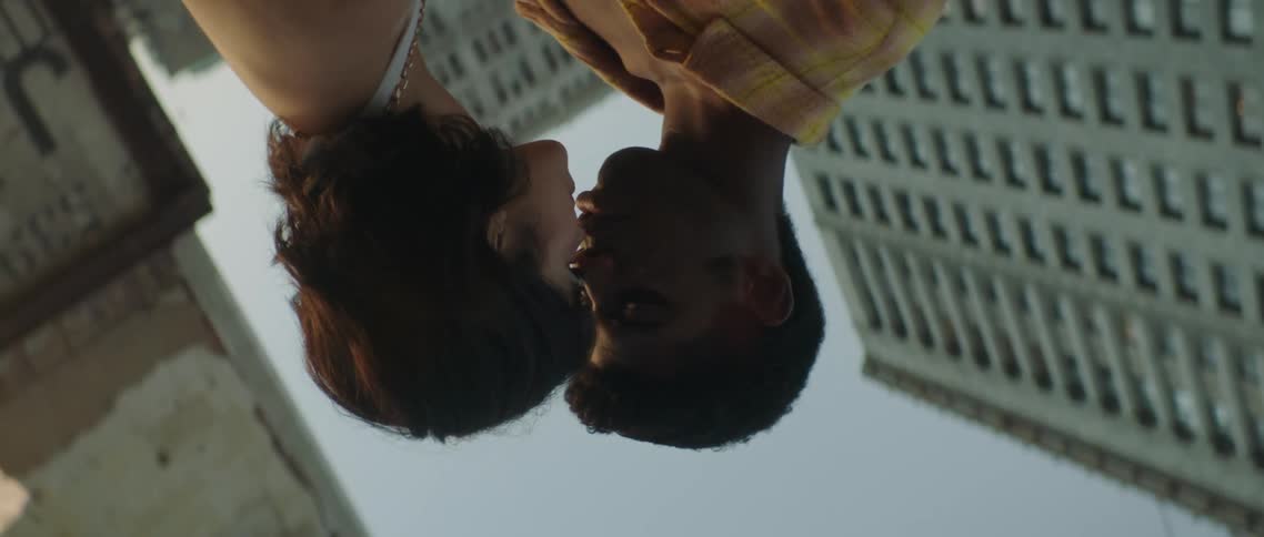 A teenage girl and boy stand on a street in front of a high rise building and kiss one another