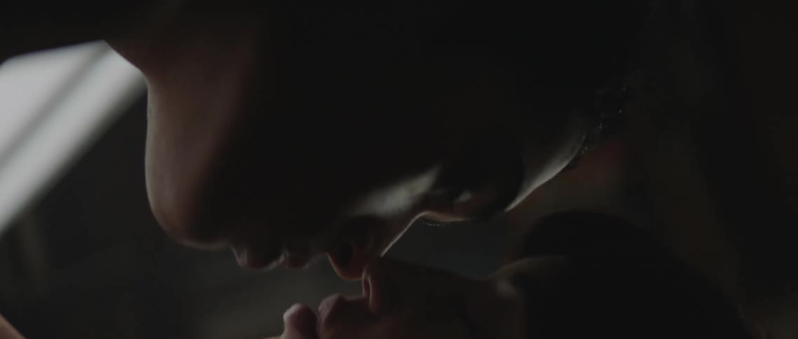 A man and a woman in a room kissing with a window and wall in the background