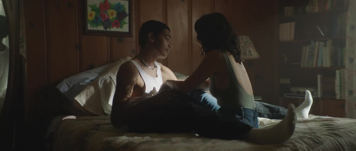 A couple sit across from each other in bed and hold hands