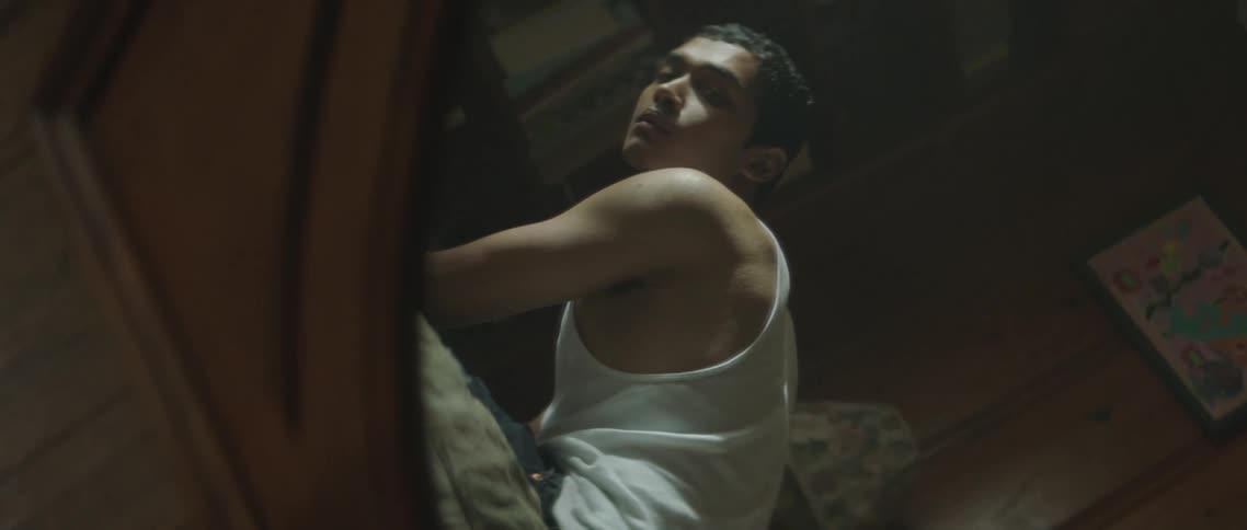 A young man wearing a white tank top sits and looks into the camera via a reflection in a nearby mirror