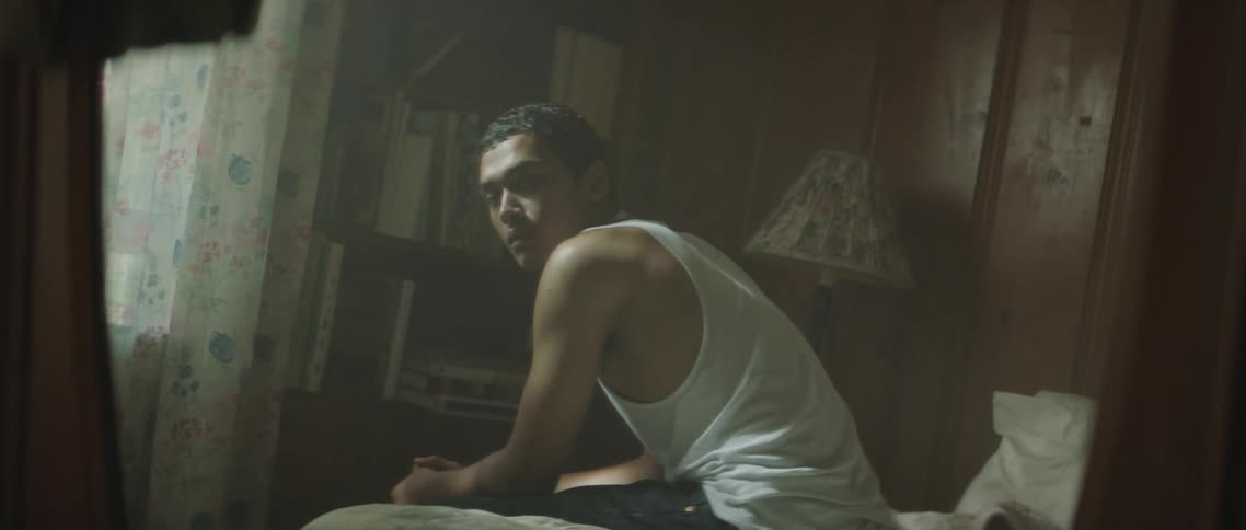 Reflection in the mirror of a young man sitting in a bed inside a dimly lit room at home