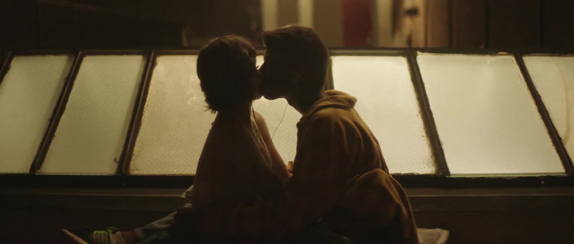Young couple hugging and kissing while sitting next to a window on the roof of a building at night