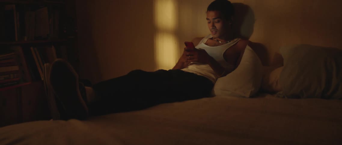 A teenage boy sits on his bed and smiles as he looks at his cell phone and texts
