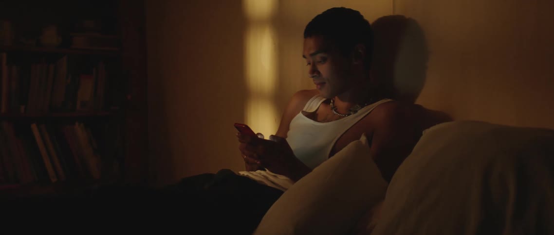 Young man sitting on bed texting on the phone inside a dimly lit bedroom