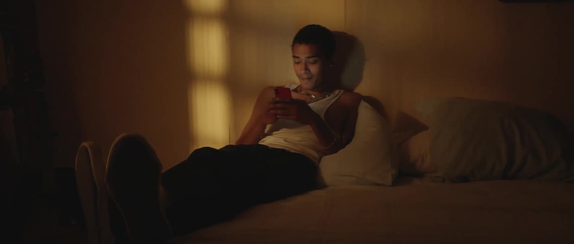 Man wearing tank top pants and shoes sits on bed in dim bedroom using mobile phone