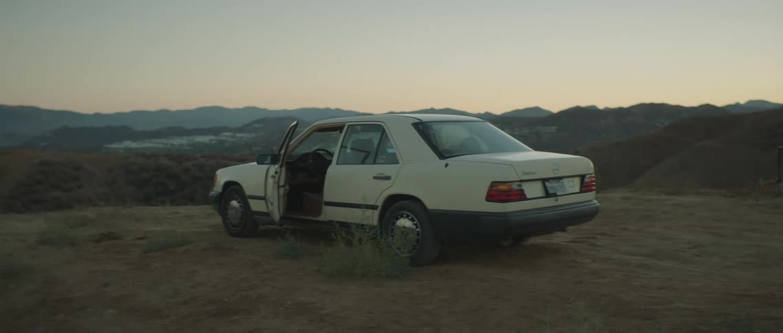 Car with open door sits parked on dirt field backed by hills and mountains stretching to pink tinted horizon