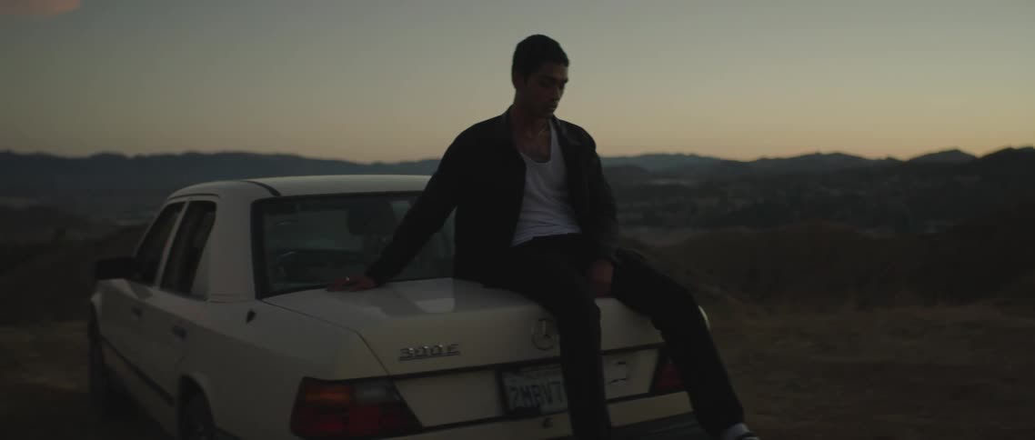 Man sits on trunk of mercedes parked on dirt area on edge of hilly terrain stretching to red tinted horizon