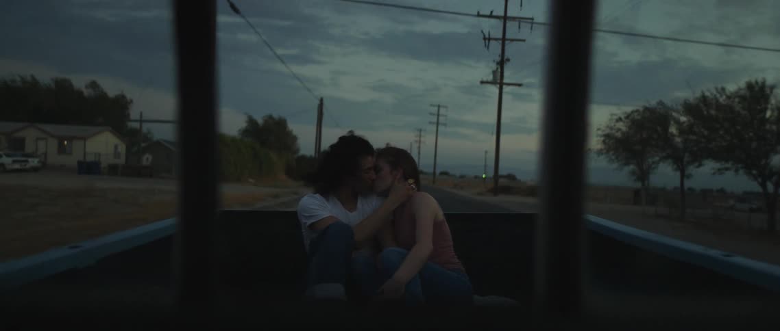 A young couple sit together and make out as they ride in the back of a pickup truck along a road at dusk