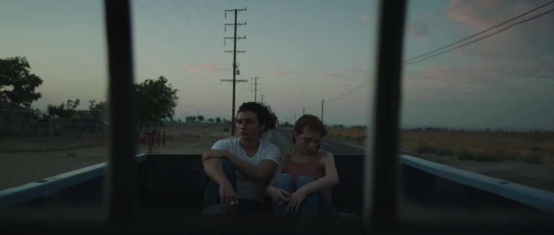 A teenage boy and girl sit in the back of a truck together as it drives down a road