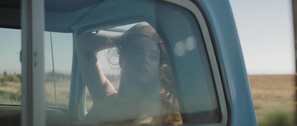 A young woman with long hair relaxes as the leans out the open window of a moving pickup truck