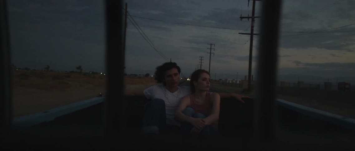 A man and woman are sitting on the open boot of a moving vehicle