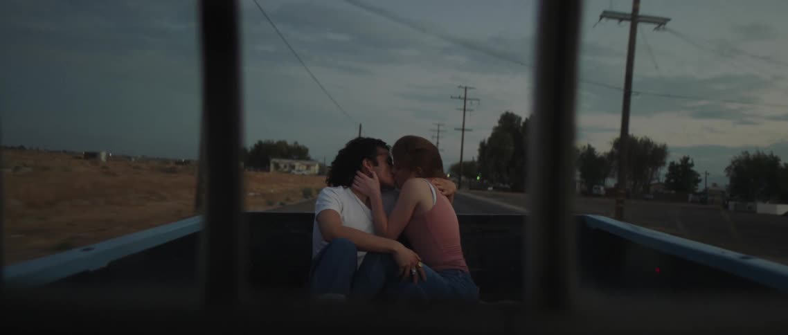 A teenage boy and girl sit in the back of a pick up truck as it drives and kiss