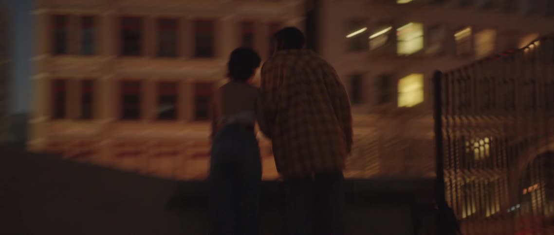A couple standing on a rooftop holds hands as they look down at a city street at night