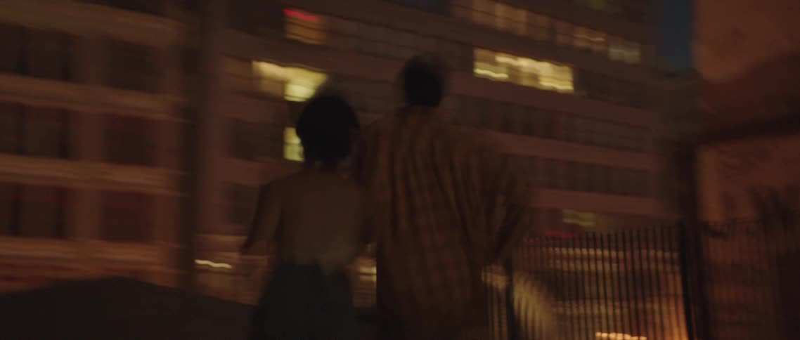 A man and woman hold hands while they run across a rooftop to the edge where they look down at the street below