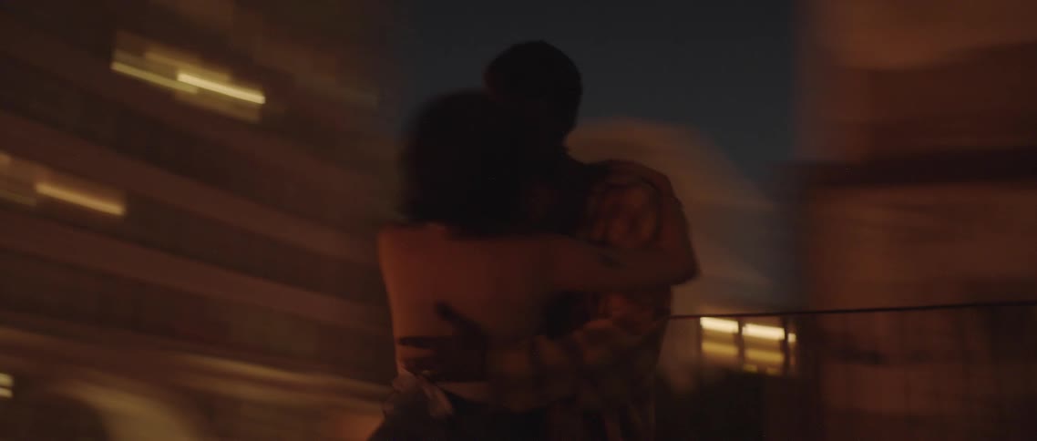 A man and a woman kissing on a rooftop with buildings and the sky and the background