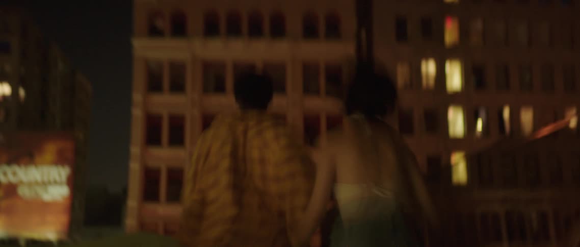 A man and woman holding hands run across a rooftop to look over the edge at the streets below
