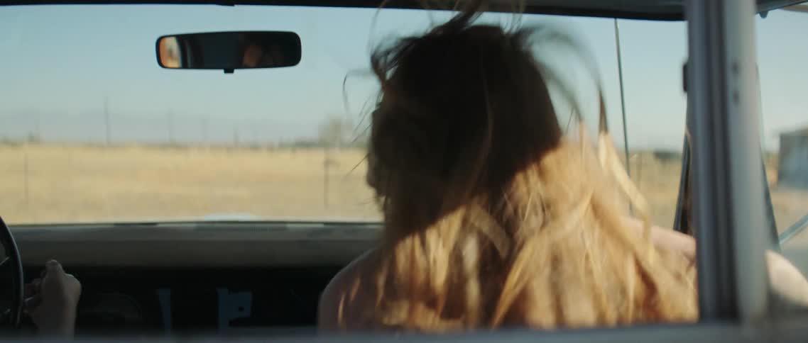 A man waits in a vehicle as a woman runs around the vehicle to climb into the passenger seat and greet him with a kiss as he begins to drive