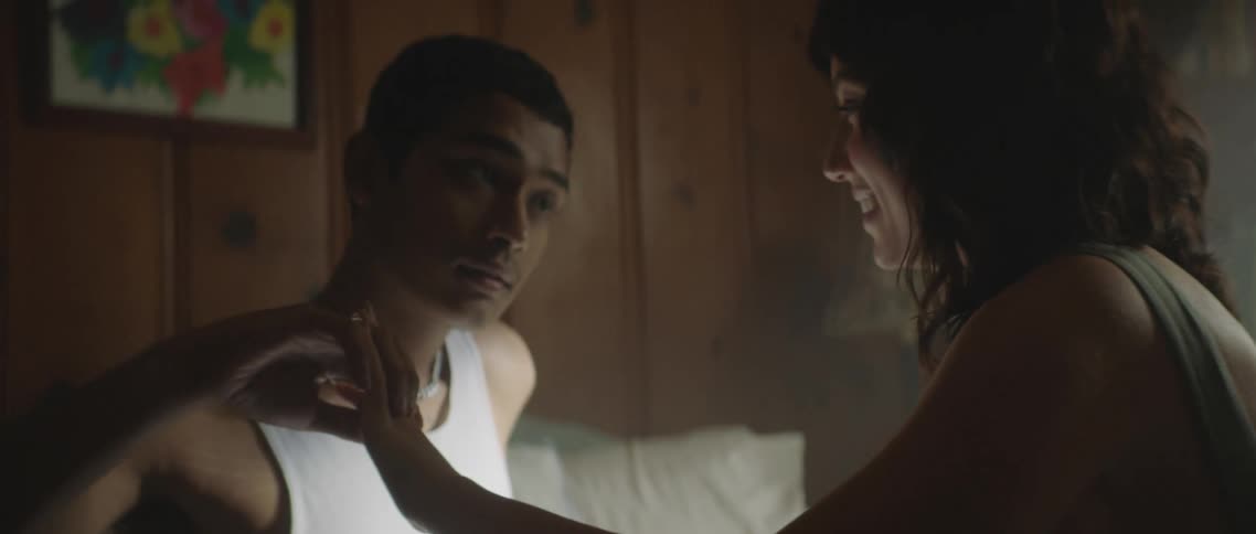 A young man and woman sit atop a bed while talking and caressing each others hands