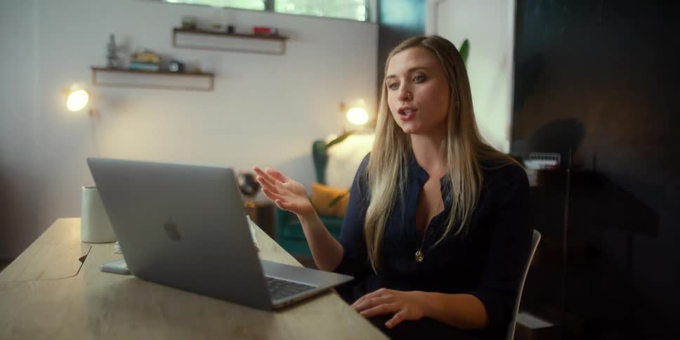 Woman sits at a desk and talks while looking at her laptop