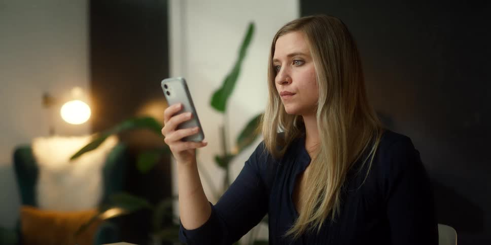 A woman in a room looking at a smartphone with a wall a lamp and door in the background