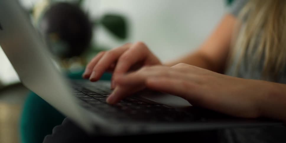 A woman in a room sitting on a couch typing on a laptop with a wall and plant in the background
