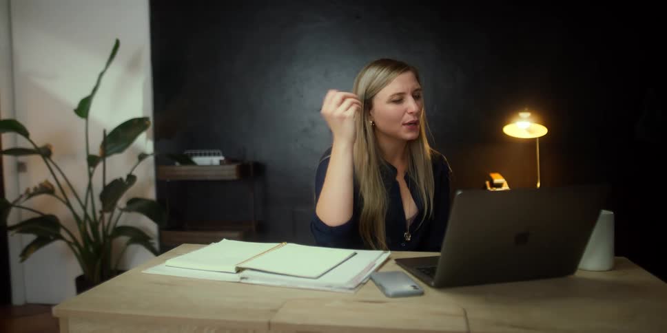A woman in a room with a pen and looking at a laptop with a wall in the background