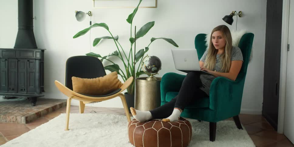 A woman typing on a laptop while sitting on a green upholstered chair in a living room