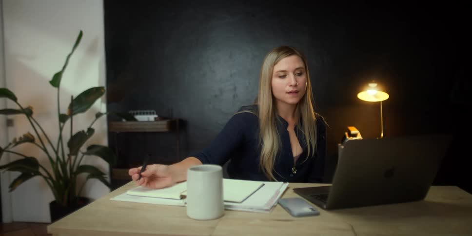 A woman in a room holding a pen and looking at a laptop with a wall in the background