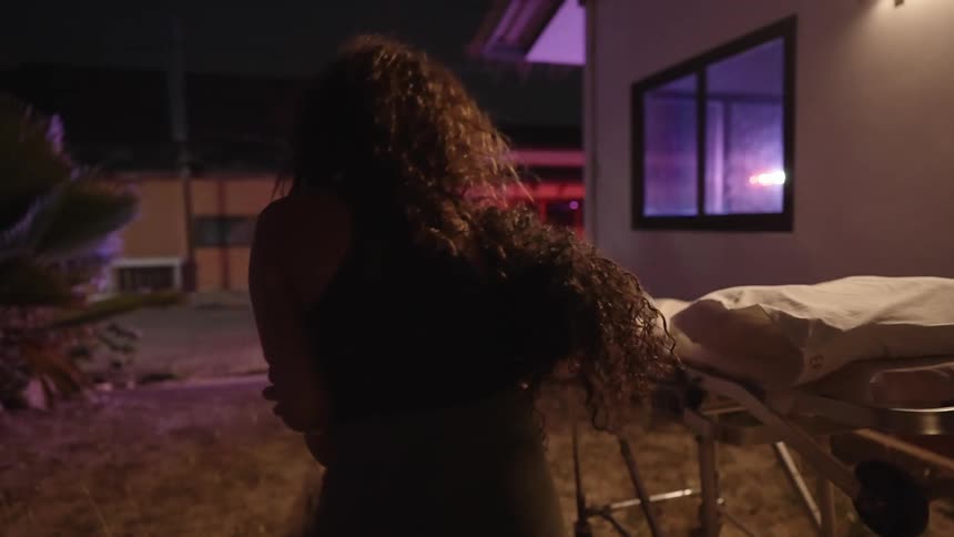 A woman holding back another woman as she cries with a stretcher buildings flashing lights and the sky and the background