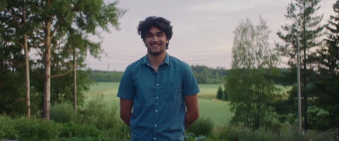 Young smiling man wearing a blue shirt smiles at the camera while standing on a field at sunset