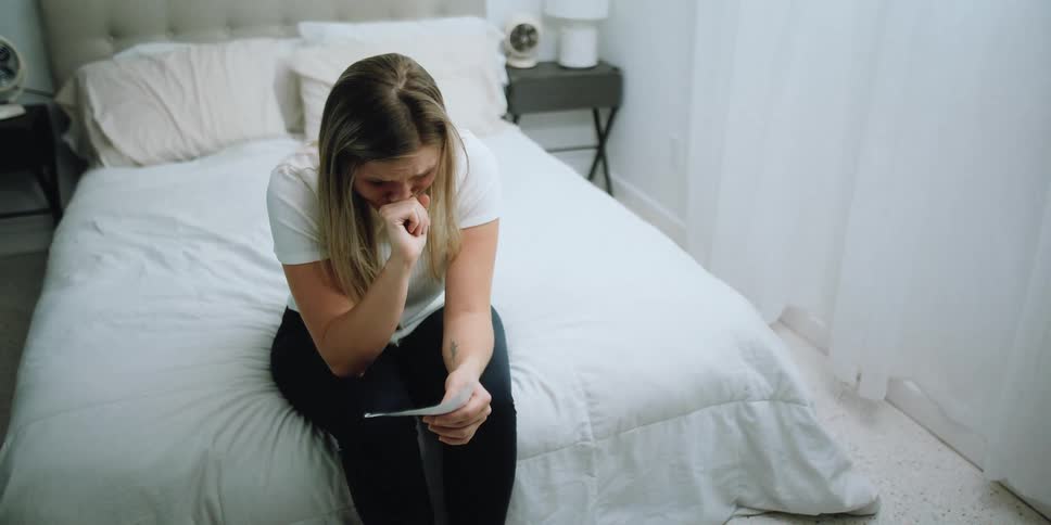 A woman sits on the end of her bed and cries while looking at a piece of paper