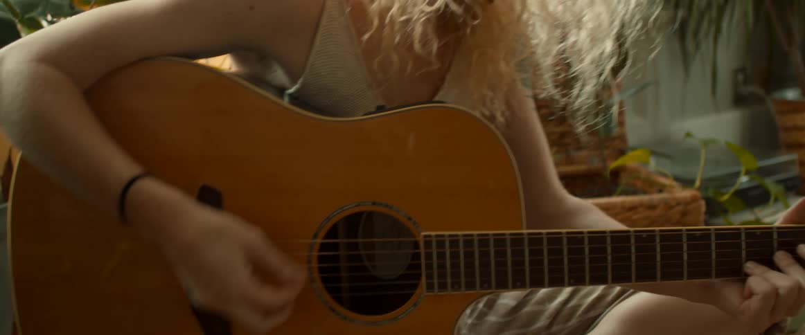 Blonde woman with curly hair sits among houseplants playing guitar
