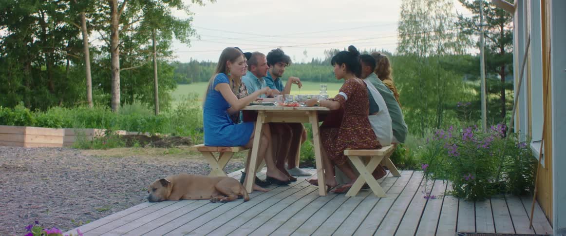 Family and friends are eating outdoors at a table on a patio with a dog relaxing nearby