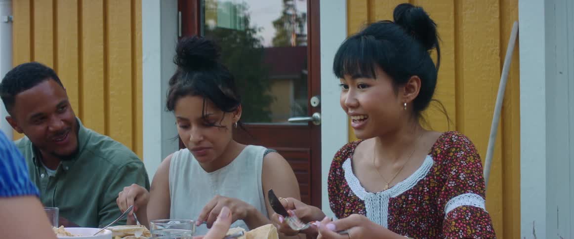 A group of friends talk and laugh as they eat together at a backyard barbecue