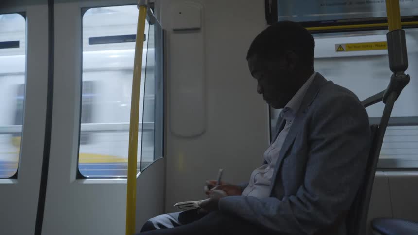 Man sits in chair on public transit writing in notepad as car passes other train