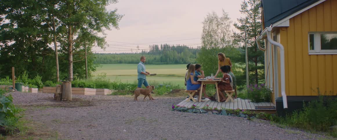 People sitting at a picnic table next to a house with trees the sky and clouds in the background