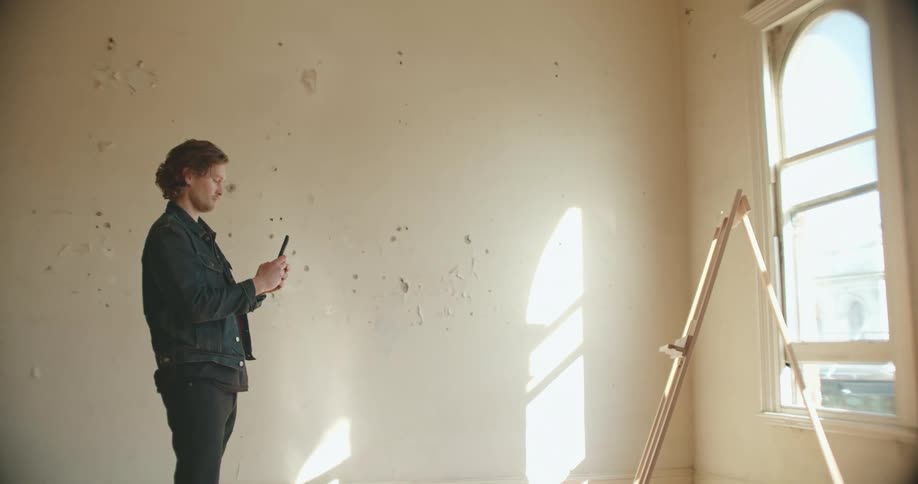 Man stands in empty room with two empty easels using mobile phone to take picture of one of the easels