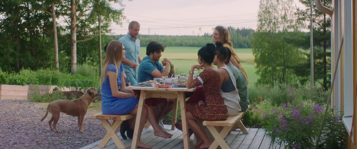 Woman in yellow dress and man with blue shirt brings food to a group of people sitting at table talking to each other in the garden