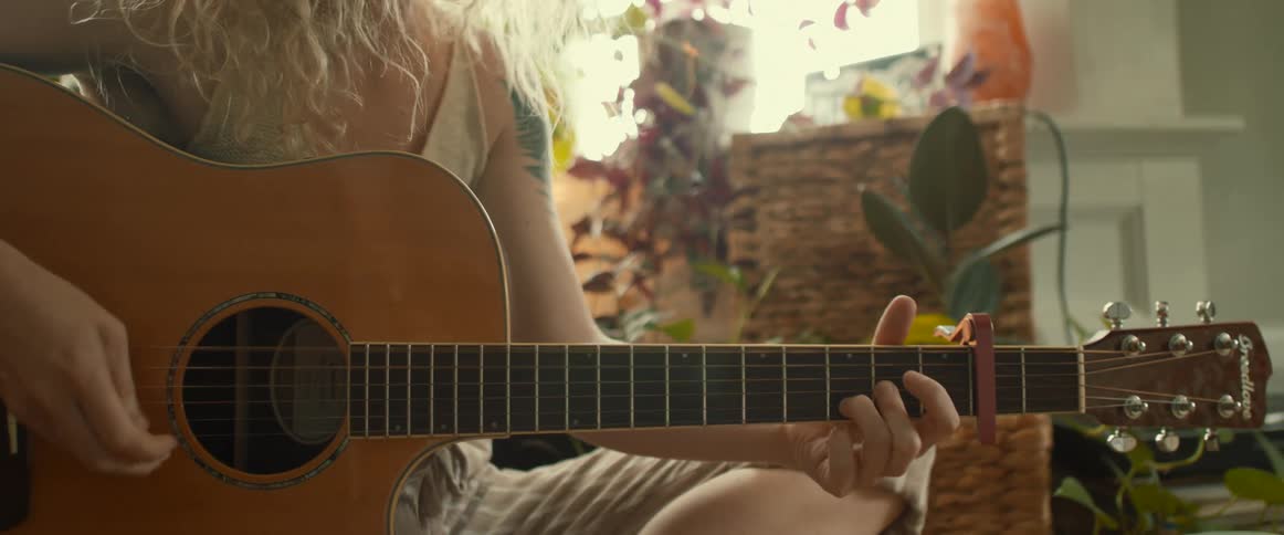 A man plays the guitar while sitting on the floor in a room filled with plants