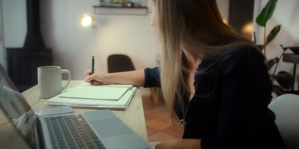 Young blonde woman takes handwritten notes while talking on a video call on a laptop
