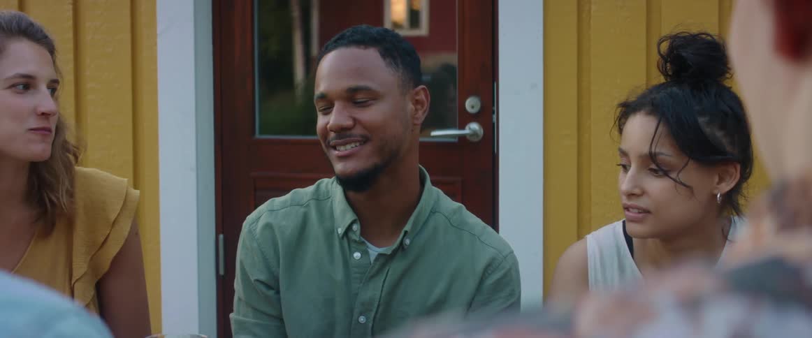 A man talks to a laughing group of friends as they sit outside eating and drinking
