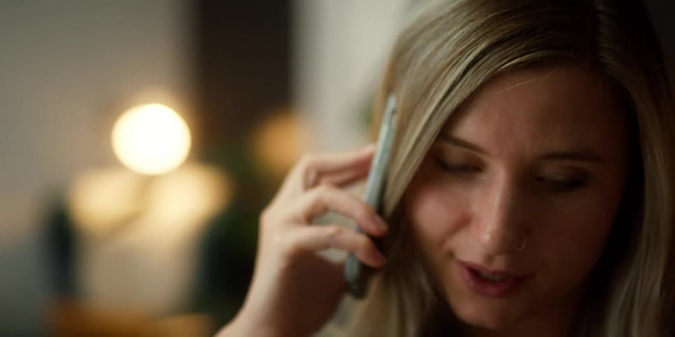 A woman working on a laptop picks up a cell phone and starts talking into it